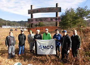 A group photo of participants holding a SUMCO flag in front of a wooden sign at the Zusayama Forest Creation activity.