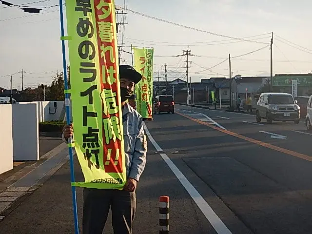 夕暮れの道路脇で、従業員が通行車両に安全運転を呼びかけている様子